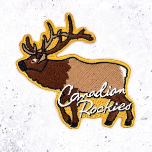 May include: Embroidered patch depicting a brown elk with large antlers, set against a tan and brown body. The patch has a yellow border and the words "Canadian Rockies" in white script.