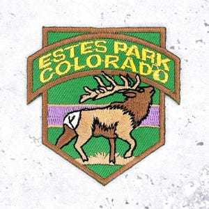 May include: Embroidered patch featuring an elk and the text "Estes Park Colorado." The patch has a green background with a brown border and a purple stripe. The elk is brown and tan.
