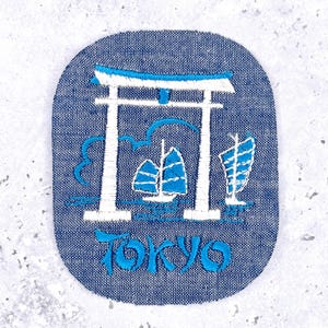 Peut inclure: Un écusson ovale en jean bleu avec une broderie blanche et bleu clair. Le motif représente un torii traditionnel, deux voiliers sur l'eau et le mot TOKYO brodé dans une police bleue stylisée au bas de l'écusson.
