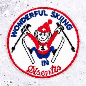 Op de afbeelding: Geborduurde patch met een rode en witte rand. De patch toont een stripfiguur skiër met een rode hoed en blauwe en rode kleding, die skistokken vasthoudt. De tekst "WONDERFUL SKIING IN Disentis" staat in een cirkelvorm.