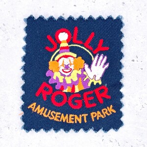 May include: Embroidered patch with a navy blue background. The patch features a colorful clown with the words "JOLLY ROGER AMUSEMENT PARK" in red and yellow. The clown has orange hair, a red nose, and a white hand waving.
