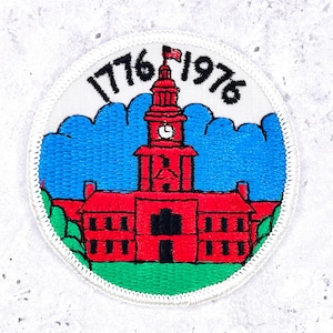 May include: A circular embroidered patch featuring a red building with a central clock tower, a small flag on top, and green ground. The text 1776 and 1976 is stitched in black above the building against a blue sky background with white borders.