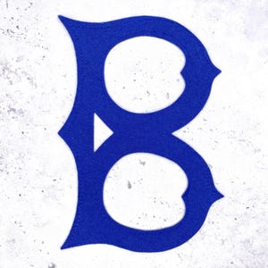 May include: A royal blue felt letter "B" with a decorative serif design. The letter is stitched with a contrasting thread and set against a textured, off-white background. The design is a bold, single-letter monogram.