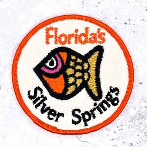 May include: Embroidered patch with a circular design featuring a stylized fish in orange, black, and gold. The text "Florida's" is above the fish, and "Silver Springs" is below, all in black lettering. The patch has an orange border.