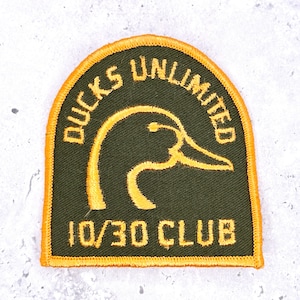 May include: A green and gold embroidered patch with the words "DUCKS UNLIMITED" arched across the top. A gold outline of a duck's head is in the center. The text "10/30 CLUB" is at the bottom. The patch has a gold border.