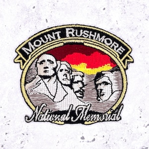 May include: Embroidered patch depicting Mount Rushmore National Memorial. The patch has a gold border with the words "Mount Rushmore" and "National Memorial" in black script. The background shows a red and yellow sunset.