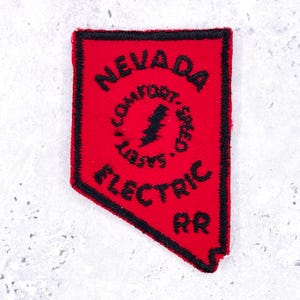 May include: A red embroidered patch shaped like the state of Nevada with a black border. The text NEVADA is at the top, ELECTRIC RR is at the bottom, and a central circle contains a lightning bolt icon with the words COMFORT, SPEED, and SAFETY.