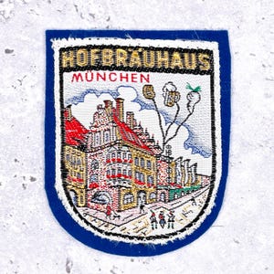 Peut inclure: Écusson brodé bleu et blanc avec le logo Hofbräuhaus München. L'écusson représente une illustration détaillée du bâtiment avec des toits rouges et le texte "HOFBRÄUHAUS MÜNCHEN" en lettres dorées. Bordure blanche.