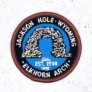 Puede incluir: Parche redondo con borde marrón, con el texto "JACKSON HOLE WYOMING" y "ELKHORN ARCH". El centro tiene un fondo azul con un diseño de arco blanco y negro y el texto "EST. 1914".