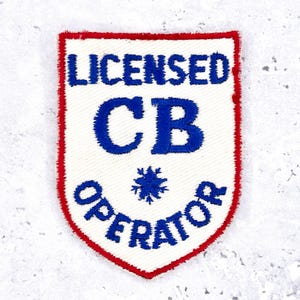 Könnte beinhalten: Ein weißes, schildförmiges Abzeichen mit rotem Rand. Das Abzeichen zeigt die Worte "LICENSED CB OPERATOR" in blauer Schrift. Ein blaues Schneeflocken-Design ist unter den Buchstaben "CB" zentriert.