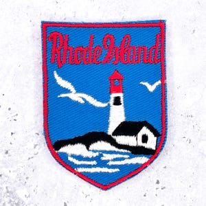May include: A shield-shaped embroidered patch with a blue background and a red border. It features the red text Rhode Island above a white lighthouse with a red top, a black roofed building, white waves, and two white seagulls in flight.