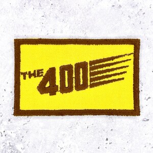 May include: A rectangular embroidered patch featuring a bright yellow background with a dark brown border. The text THE 400 is stitched in dark brown, with the number 400 featuring horizontal motion lines extending to the right side of the patch.