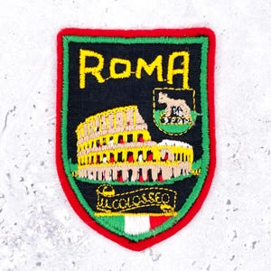 May include: A shield-shaped embroidered patch with a black background, a red border, and a green inner trim. It features the yellow text ROMA, an illustration of the Colosseum, a small emblem with a wolf, and the text IL COLOSSEO above an Italian flag.