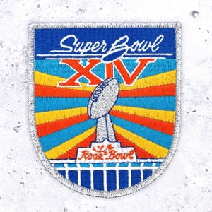 Könnte beinhalten: Gestickter Aufnäher mit den Worten "Super Bowl XIV" in weißer Schrift und römischen Ziffern. Der Aufnäher hat einen blauen Rand und ein Design einer Football-Trophäe und eines Footballs. Der Hintergrund hat ein Streifenmuster in Orange, Gelb und Blau.