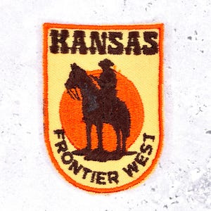 Peut inclure: Écusson brodé en forme de bouclier, avec le texte "KANSAS" en haut et "FRONTIER WEST" en bas. Une silhouette marron d'un cow-boy à cheval est centrée sur un soleil orange.
