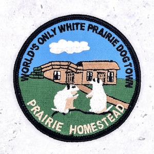 May include: A circular embroidered patch with a black border featuring a light blue sky, a white cloud, and a tan building. Two white prairie dogs stand on green grass. The text reads: WORLD'S ONLY WHITE PRAIRIE DOGTOWN and PRAIRIE HOMESTEAD.