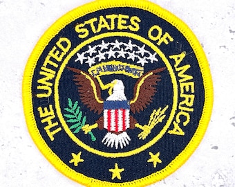 May include: Circular patch with a yellow border and the words "THE UNITED STATES OF AMERICA" in yellow. The patch features a blue background with a central emblem of an eagle, stars, and text.