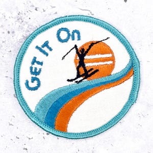 May include: A circular embroidered patch with a white background and a teal border. It features the blue text "Get It On" above a black silhouette of a skier on an orange sun. Below, there are two curved stripes in teal and orange.