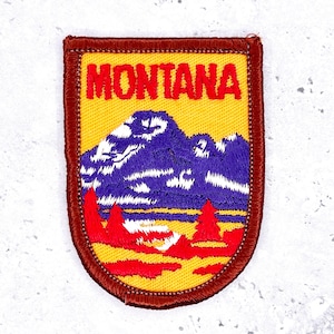 May include: Embroidered patch featuring the word "MONTANA" in red block letters above a mountain scene. The patch has a yellow background with a purple mountain range and red trees. A brown border frames the design.