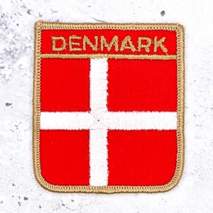 May include: A rectangular embroidered patch featuring the flag of Denmark with a bright red background and a white cross. The word DENMARK is stitched in gold thread at the top, and the entire patch is bordered by a thick gold embroidered edge.
