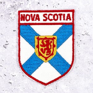 May include: Embroidered patch featuring the Nova Scotia flag. The patch has a white background with a blue St. Andrew's cross, a red border, and a gold and red shield in the center. The text "NOVA SCOTIA" is at the top.