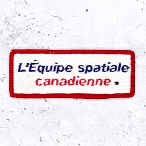 Peut inclure: Un écusson rectangulaire brodé sur fond blanc avec une bordure rouge. Le texte L'Équipe spatiale est brodé en bleu foncé, tandis que le mot canadienne est en rouge, accompagné d'une petite étoile noire sur le côté droit de l'écusson.