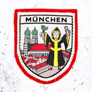 May include: A shield-shaped embroidered patch with a red border featuring the text MÜNCHEN at the top. The design depicts a figure in a black and yellow robe holding a green plant and a mug, standing before a city scene with red-roofed buildings.