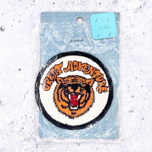 May include: Round patch with a tiger design and the words "GREAT ADVENTURE" in orange lettering. The tiger is orange and black with a red mouth. The patch is on a light blue backing.