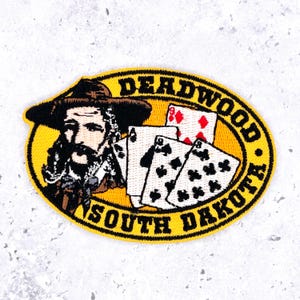 Op de afbeelding: Een ovale geborduurde patch met een gele achtergrond met een man met een hoed met brede rand, vier speelkaarten en de zwarte tekst DEADWOOD SOUTH DAKOTA. De man heeft een donkere baard en snor, en de kaarten tonen verschillende symbolen.