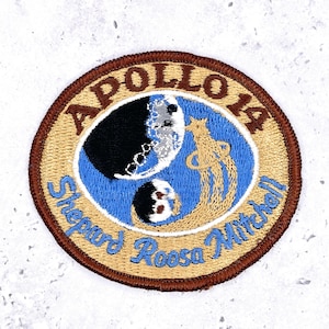 May include: A circular embroidered patch featuring the Apollo 14 mission emblem. The design includes a blue center with a black and white moon, a gold star, and gold trail. Text reads APOLLO 14 and Shepard Roosa Mitchell in brown and blue.