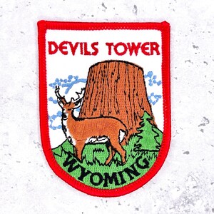 May include: Embroidered patch featuring Devils Tower in Wyoming. The patch has a red border and includes a brown tower, a deer, and the text "DEVILS TOWER WYOMING" in red and green.