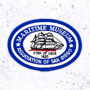 May include: An oval patch with a blue border and white interior. The patch features the text "MARITIME MUSEUM ASSOCIATION OF SAN DIEGO" and a black and red illustration of a tall ship. The text "STAR OF INDIA 1863" is also present.