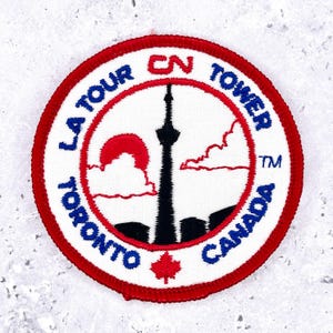 May include: Embroidered patch featuring the CN Tower in Toronto, Canada. The design includes the tower silhouette, a red sun, and the text "LA TOUR TORONTO CANADA" in blue, with a red border. A maple leaf is at the bottom.