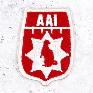 May include: A shield-shaped embroidered patch featuring a red background with a white border. The text AAI is stitched in white at the top. Below the text, a white eight-pointed star contains a red silhouette of a standing animal figure.