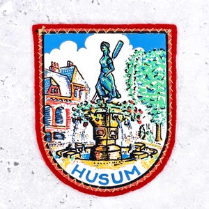 May include: A shield-shaped embroidered patch with a red border featuring a colourful illustration of a fountain with a central figure, a building, and a tree under a blue sky. The word HUSUM is printed in blue capital letters at the bottom.