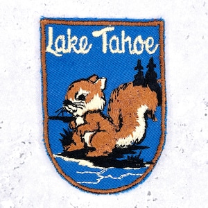 May include: A shield-shaped embroidered patch featuring a blue background with the text Lake Tahoe in white script. The design includes a brown squirrel with a bushy tail, black silhouettes of pine trees, and a white shoreline detail at the base.