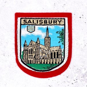 May include: Embroidered patch featuring a detailed illustration of Salisbury Cathedral, set against a blue sky with clouds. The patch has a white border, a red frame, and the word "SALISBURY" at the top.
