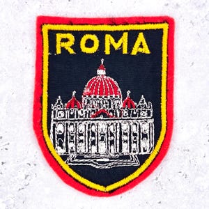 May include: A shield-shaped embroidered patch with a black background and a red border. The word ROMA is stitched in yellow at the top. Below the text, a white and red embroidered depiction of a domed building is centered on the patch.
