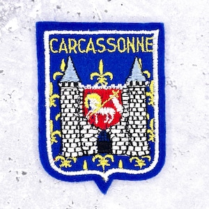 May include: A shield-shaped embroidered patch featuring a blue background with a white border. The text CARCASSONNE is stitched in yellow at the top. It depicts a white stone castle with two towers and a red central shield with a white horse.