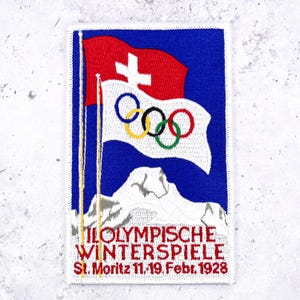 May include: A rectangular embroidered patch featuring a blue background with a red Swiss flag and a white Olympic flag with five rings. Below, a white mountain peak sits above the red text II. OLYMPISCHE WINTERSPIELE St. Moritz 11.-19. Febr. 1928.