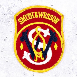 May include: A shield-shaped embroidered patch with a red background and yellow border. The text SMITH & WESSON is stitched in yellow at the top. The center features a large white S intertwined with a dark blue W and a yellow decorative flourish.