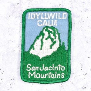 May include: A rectangular embroidered patch with a green border. The design features a light blue sky above a white mountain peak and green foreground. The text reads IDYLLWILD CALIF at the top and San Jacinto Mountains at the bottom in white.