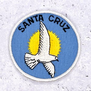 May include: Embroidered patch featuring a white bird in flight against a yellow sunburst on a light blue background. The words "SANTA CRUZ" are arched around the top edge in black lettering. The patch has a white stitched border.