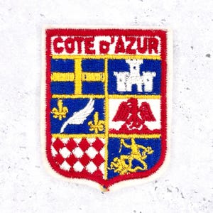 May include: A rectangular patch with a red border and the words "COTE D'AZUR" in red letters. The patch features a blue and yellow cross, a white castle, a white feather, a red eagle, and a yellow figure.