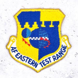 May include: Embroidered patch with a shield design. The shield is divided into yellow and blue sections, featuring a globe, stars, and the text "AF EASTERN TEST RANGE". The patch has a yellow border.
