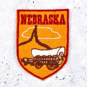 May include: Embroidered patch featuring the word "NEBRASKA" in red block letters above a brown and white covered wagon and a brown ox. The patch has a gold background and a red border.