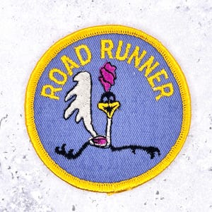 May include: A circular embroidered patch featuring a light blue background with a yellow border. The text ROAD RUNNER is stitched in yellow letters at the top. A stylized bird graphic with a white wing, pink crest, and yellow beak is centered.