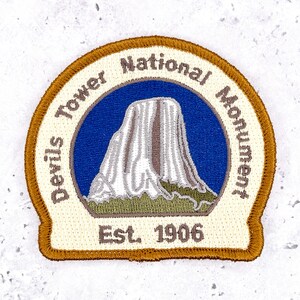 May include: A circular embroidered patch featuring a depiction of Devils Tower against a blue sky and green base. The border is gold, and the cream-colored background displays the text Devils Tower National Monument and Est. 1906 in brown.