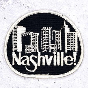 Op de afbeelding: Een zwart-wit geborduurde patch met een silhouet van een stad en het woord "Nashville!" in witte letters. De patch heeft een witte rand en is bedoeld voor kleding of accessoires.