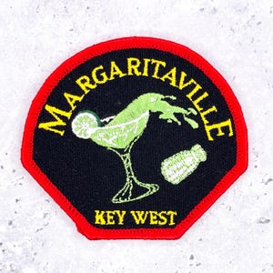 May include: A black embroidered patch with a red border featuring a lime green margarita glass, a salt shaker, and the yellow text MARGARITAVILLE and KEY WEST. The design includes a lime slice on the glass rim and a splash of liquid.
